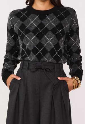 Kvinde iført sort og grå argyle sweater stoppet ned i højtaljede kulstofgrå bukser med bundet bælte, hænderne i lommerne og guld armbånd på håndleddet.