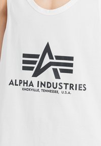 Bela brezrokavna majica s črnim grafičnim logotipom, ki vključuje dve vodoravni črti in besedilo "ALPHA INDUSTRIES" ter podrobnosti o lokaciji spodaj.