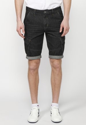 Homme portant un short cargo en denim noir avec des revers retroussés et des baskets blanches, debout devant un fond uni.