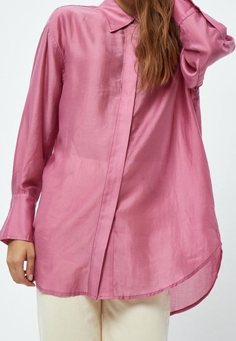 Femme portant une chemise rose ample à manches longues avec col et boutons, associée à un pantalon clair, posant avec une main levée.