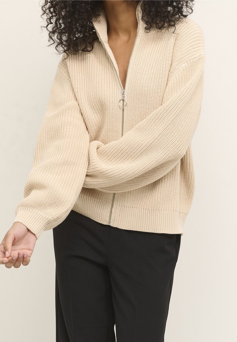 Personne portant un pull zippé en maille épaisse beige avec des manches bouffantes et un pantalon noir, debout devant un fond clair uni.