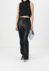 Top negro corto sin mangas con lazos de tela, combinado con pantalones de cuero negros de cintura alta. Sosteniendo un bolso plateado anudado.