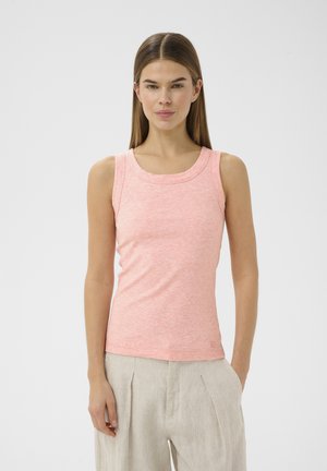 Jeune femme aux cheveux bruns raides, portant un débardeur rose côtelé et un pantalon en lin beige, debout avec une main dans la poche.