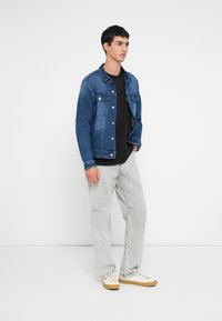 Junger Mann steht, trägt eine blaue Jeansjacke, ein schwarzes Hemd, hellgraue Jeans und weiße Turnschuhe mit Gummisohlen vor einem schlichten weißen Hintergrund.