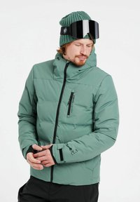 Grüne Pufferjacke mit einem Reißverschluss-Brusttasche, verstellbaren Bündchen und Kapuze. Getragen mit einer Strickmütze und großen schwarzen Skibrillen.