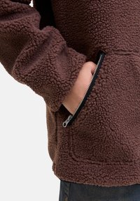 Hnedá fleecová bunda s textúrovaným, nadýchaným povrchom. Má bočnú vrecko na zips a elastické manžety pre tesné uchytenie.