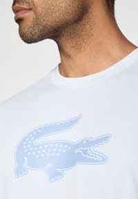 Balta kokvilnas T-krekls ar lielu gaiši zilu krokodila logotipu ar ģeometriskām raksturojumiem, apaļu kaklu un īsām piedurknēm.