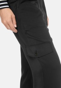 Schwarze Cargo-Hosen mit einer Klappentasche mit Knopf, glatter Textur und figurbetontem Design. Sichtbare Handposition auf dem Oberschenkel.