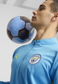 Lichtblauwe sportjacket met een ritssluiting bij de kraag, voorzien van een Manchester City-logo en een gele Puma-accent, met een zwart en blauwe voetbal in de buurt.