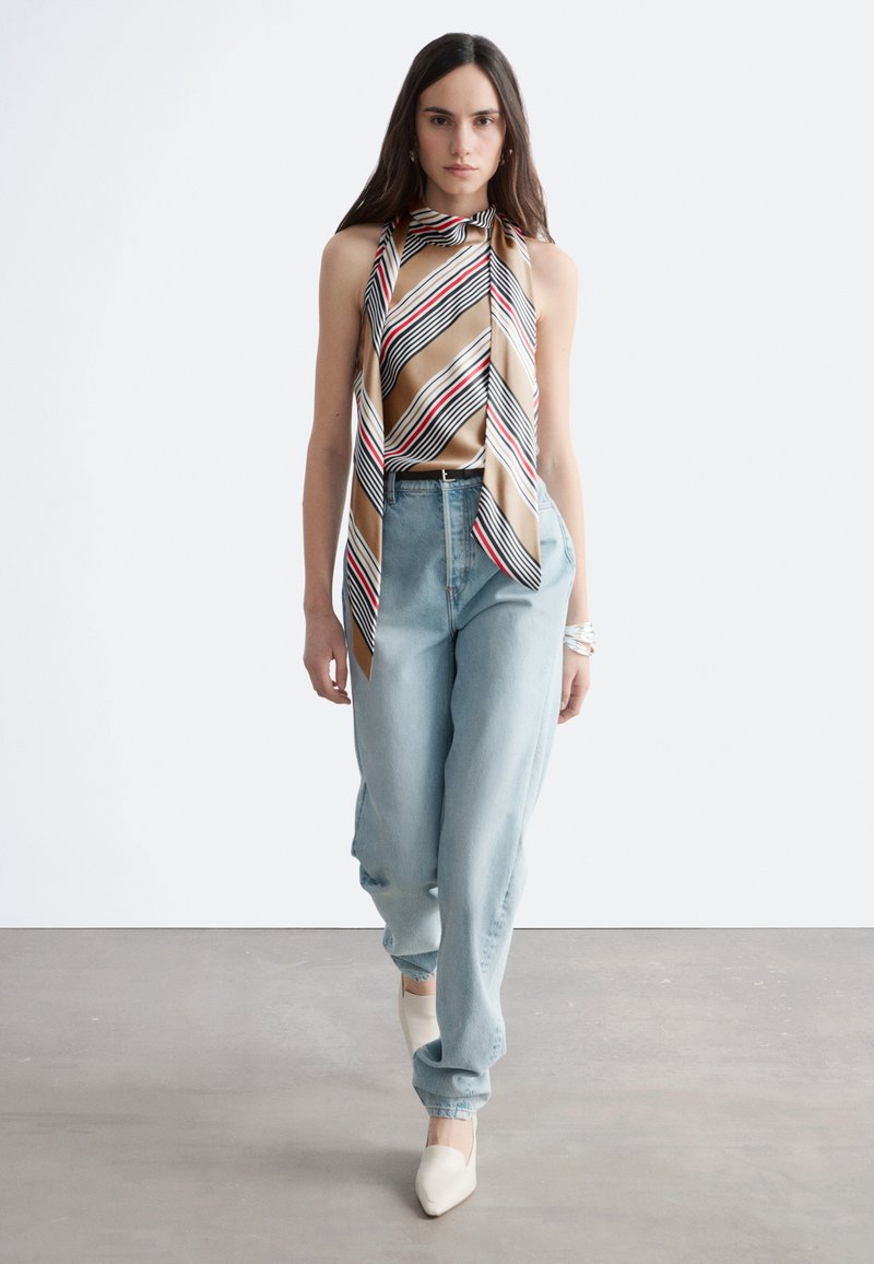 Femme marchant vers l'avant portant un haut sans manches à rayures beige en forme d'écharpe, un jean taille haute bleu clair, des chaussures blanches pointues et des bracelets argentés.