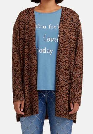 Kvinde iført en brun cardigan med leopardprint over en blå skjorte med teksten "Du føler dig elsket i dag" og blå jeans med diamantmønster.