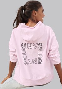 Felpa rosa con vestibilità rilassata, cappuccio e grande testo nero stampato sul retro. Il tessuto sembra morbido e ha una texture liscia.