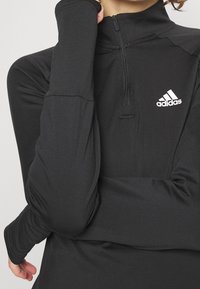 Schwarzer Halbzip-Pullover aus glattem Material, mit Daumenlöchern, einem hohen Kragen und einem weißen Adidas-Logo auf der linken Brust.