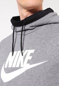 Šedá mikina s kapucí a černým lemováním u krku, zdobená výrazným bílým logem Nike a černými stahovacími šňůrkami. Měkká textura a sportovní střih.