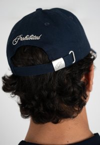 Persona con una gorra azul marino puesta al revés con la palabra "Prohibido" bordada en blanco y una hebilla ajustable plateada en la correa.