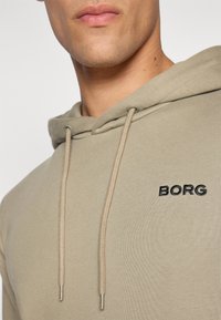 Olíva zöld kapucnis pulóver, állítható zsákfüllel, fekete "BORG" hímzéssel a mellrészen és puha, texturált anyagú felülettel.