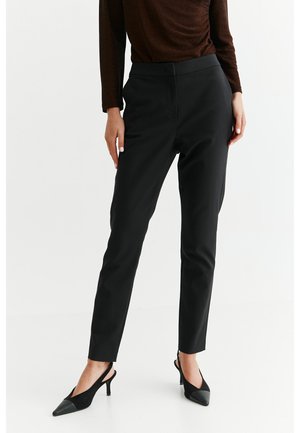 Vrouw draagt strakke zwarte broek, bruine top met lange mouwen en zwarte puntige slingback hakken, staand tegen een witte achtergrond.