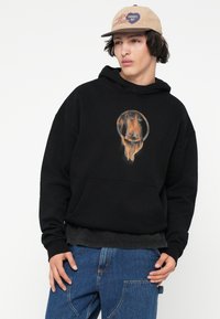 Jovem vestindo um boné bege, hoodie preto com um design de smiley face a pingar, e jeans azuis, posando contra um fundo branco.