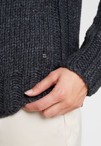 Tmavě šedý žebrovaný pletený svetr z husté příze, s malým černým knoflíkem poblíž lemu a texturovaným povrchem.