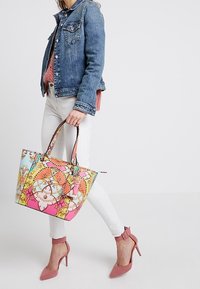 Kleurrijke tote bag met een mix van oranje, roze en blauwe patronen; heeft twee handvatten, een decoratieve hanger en een gestructureerde rechthoekige vorm.