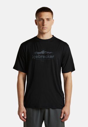 Mann mit kurzen Haaren, der ein schwarzes Icebreaker T-Shirt mit Bergmotiv und graue Hose trägt und vor einem einfarbigen weißen Hintergrund steht.