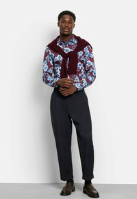 Blouse met knoopsluiting in een blauw bloemenpatroon op een bordeauxrode achtergrond, gecombineerd met donkere broeken. Trui over de schouders gedrapeerd, bruine schoenen.