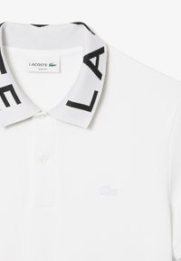 Vit slim-fit pikétröja med en texturerad krage som har svarta geometriska mönster och en liten broderad lila Lacoste-logotyp på bröstet.