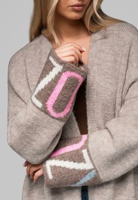 Cardigan beige chiaro con una texture morbida, caratterizzato da motivi grafici audaci in rosa, bianco e marrone sui polsini. Design con apertura frontale.