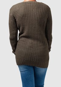 Personne portant un pull en tricot texturé marron foncé et un jean bleu, vue de dos sur un fond clair uni.