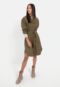 Olivgrünes Hemdkleid mit Gürtel, langen Ärmeln, Knopfleiste und seitlichen Schlitzen. Kombiniert mit beigen Schnürstiefeletten.