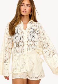 Jeune femme portant un haut boutonné en dentelle crochet crème et un short assorti, aux cheveux longs et boucles d'oreilles anneaux dorés, sur un fond blanc.