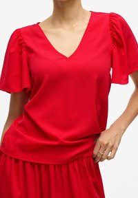 Czerwona bluzka z dekoltem w serek i krótkimi bufiastymi rękawami. Miękka, gładka tkanina; luźny krój z elastycznym pasem. Prosty wzór, bez wzorów.