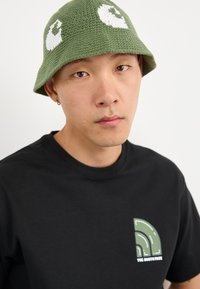 Ung person iført en grøn strikbucket hat med hvide mønstre og en sort T-shirt med et grønt og hvidt "The North Face" logo.