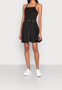 Svart klänning med figurnära överdel, fyrkantig halsringning och tunna axelband. Utsvängd kjol. Har en midja med Calvin Klein-logga. Parat med vita sneakers.