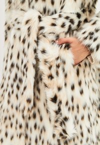 Krém színű, sötétbarna foltokkal ellátott műszőrme kabát. Övvel ellátott derék és puha, bolyhos textúra jellemzi. A kabátot egy kéz tartja.