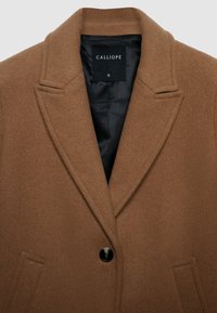 Cappotto in misto lana marrone con colletto a tromba, chiusura con un singolo bottone nero e tasche laterali. Interno foderato in nero. Taglia S.