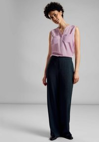 Blusa senza maniche di un leggero viola con tasche frontali, abbinata a pantaloni a gamba wide textured neri e scarpe nere. Design semplice e pulito.