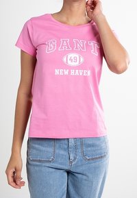 Žena nosící růžové tričko GANT s textem "49" a "New Haven", doplněné světle modrými džíny, na bílém pozadí.