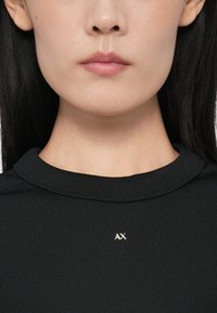 Κοντινό πλάνο του κάτω προσώπου και του λαιμού ενός ατόμου που φοράει ένα μαύρο ριμπ πουκάμισο με ένα μικρό μεταλλικό λογότυπο "AX" στο στήθος.