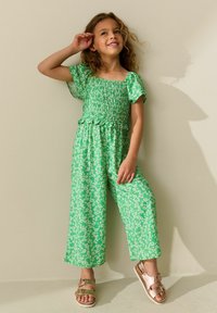 Zöld virágmintás overál rövid ujjakkal, ráncolt felsőrésszel és bő szárakkal. Csillogó aranyszandállal párosítva. Könnyű textúra és laza szabás.