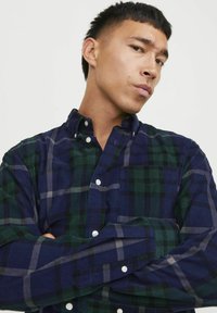 Camisa de cuadros en colores azul marino y verde con mangas largas, que presenta líneas grises en contraste y botones blancos. Tela de textura suave.