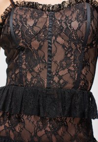 Černý krajkový top s volánkovým dekoltem a vrstveným peplum lemem. Materiál je průsvitný s květinovými vzory a saténovými akcenty podél švů.