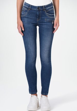 Frau trägt eine eng anliegende blaue Jeans mit Reißverschlusstasche vorne und weißen Sneakern, steht vor einem schlichten hellen Hintergrund.