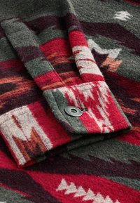 Tissu en laine plié avec un motif géométrique rouge, gris et blanc et un bouton-pression en métal au niveau du poignet.