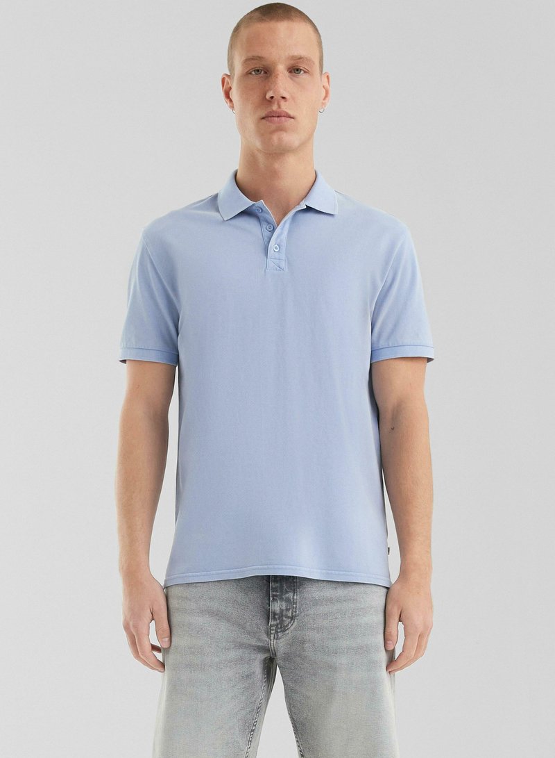 Polo shirt bleu clair avec une patte de boutonnage à trois boutons, manches courtes et texture douce ; associé à un jean gris clair.