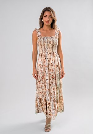 Femme portant une robe longue sans manches à motifs avec bretelles volantées et des sandales beiges, debout devant un fond gris clair uni.