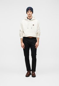 Κρεμ φούτερ με κουκούλα και λογότυπο, μαύρο παντελόνι, καφέ παπούτσια και σκούφο πλεκτό σκούρου μπλε χρώματος. Καθημερινό ντύσιμο με μινιμαλιστικό σχέδιο και ουδέτερα χρώματα.