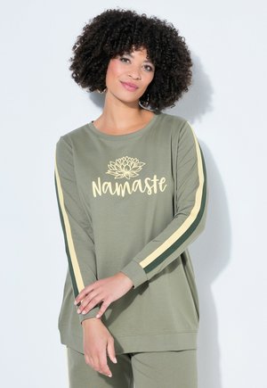 Kobieta o kręconych włosach, ubrana w oliwkowozieloną bluzkę z długim rękawem, z żółtymi i ciemnozielonymi paskami oraz napisem "Namaste" i grafiką kwiatu lotosu.