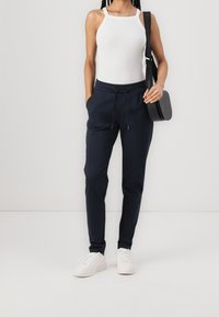 Mörkblå joggingbyxor med elastisk midja och dragsko, kombinerade med en vit ärmlös topp och vita sneakers. Svart axelremsväska.