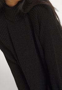 Pull en tricot noir avec un col montant côtelé et un tissu texturé. Manches longues avec une coupe décontractée, mettant en valeur un design simple.